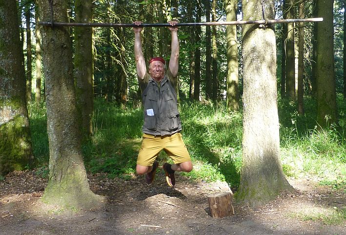 Man jumping and gripping a horizontal bar between trees in a forest clearing.